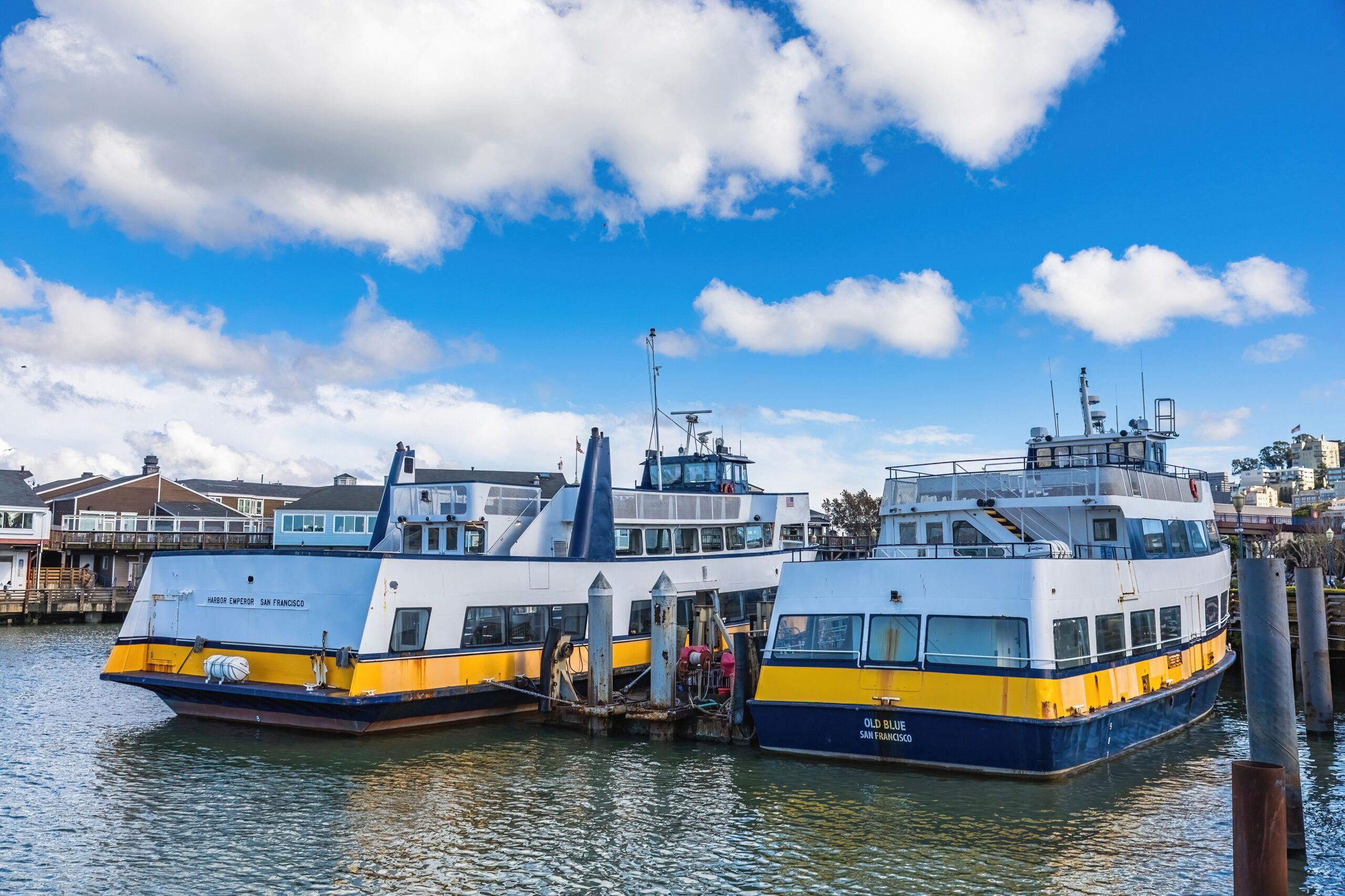 Two blue and yellow passenger ferries docked on San Francisco Bay under a blue sky with white clouds